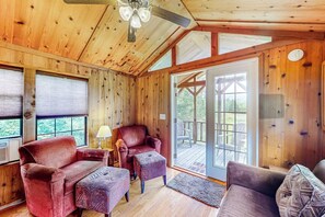 A wooden cabin interior with a ceiling fan, a red leather chair, a patterned ottoman, and a view through a glass door to an outdoor deck.
