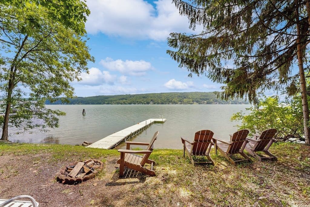 A lakeside view with a dock extending into the water, surrounded by trees and wooden chairs.