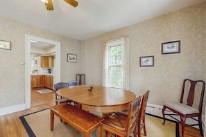 Un coin repas avec une table ronde, des chaises en bois et un banc. Il y a une fenêtre avec des rideaux, une porte donnant sur la cuisine et des tableaux encadrés accrochés au mur.
