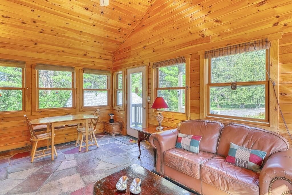 A wooden cabin interior with a dining table, leather sofa, and large windows.