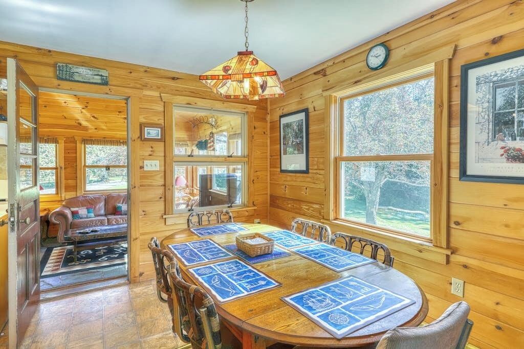 Interior de una cabaña de madera con una mesa de comedor, sillas, una araña y ventanas amplias.
