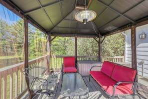 A covered outdoor seating area with a table and two red cushioned chairs, surrounded by a wooden deck and a metal railing. - 4BR Lakefront Adirondacks - Dock (Brant Lake)