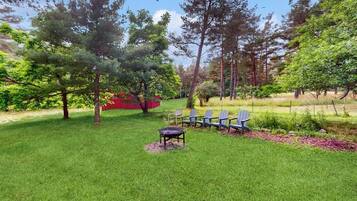 Un espace extérieur verdoyant, avec un foyer et des chaises, entouré d’arbres.