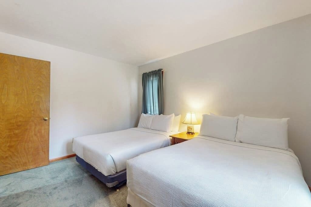 Ein Hotelzimmer mit zwei Betten, einer Holztür, einem Fenster mit Vorhängen und einem Nachttisch mit Lampe.