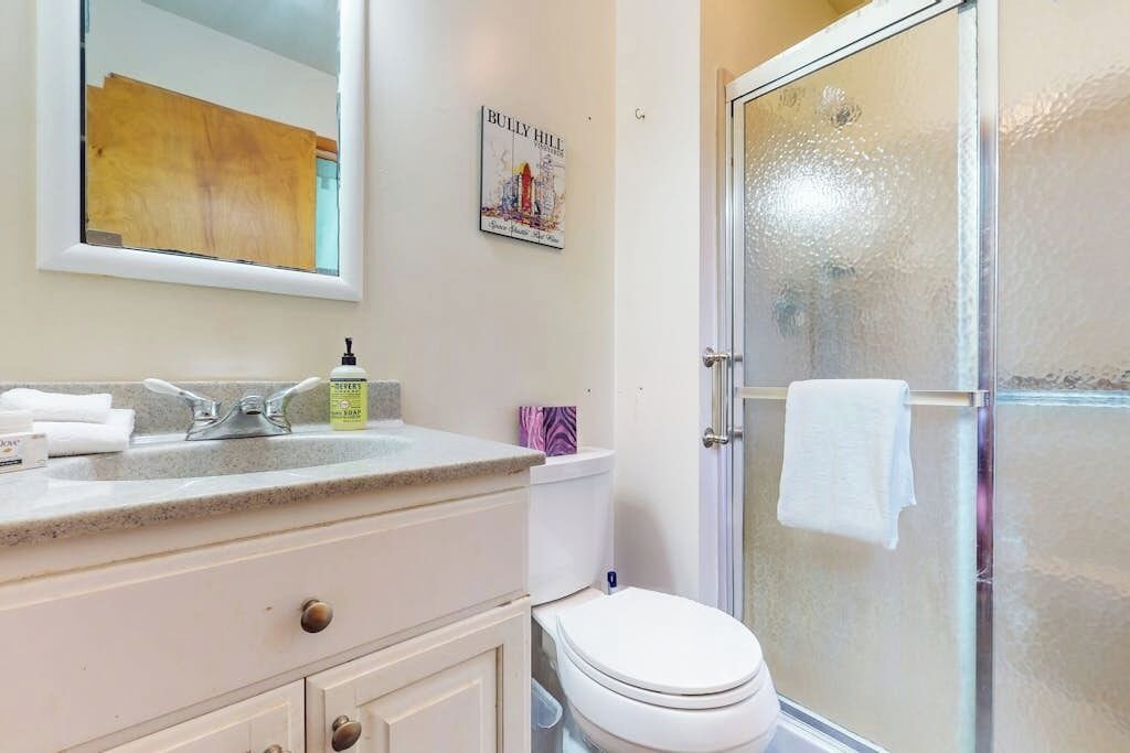 Un baño con lavamanos blanco, espejo y retrete, una ducha con puerta de vidrio y un cuadro en la pared.