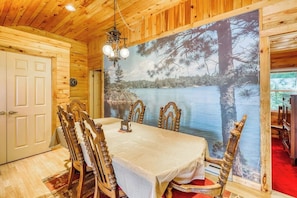 A dining area with a table set for four, wooden chairs, a chandelier, and a large wall mural of a lake and trees. - Peaceful 3BR Lakefront Adirondacks | Dock (Brant Lake)