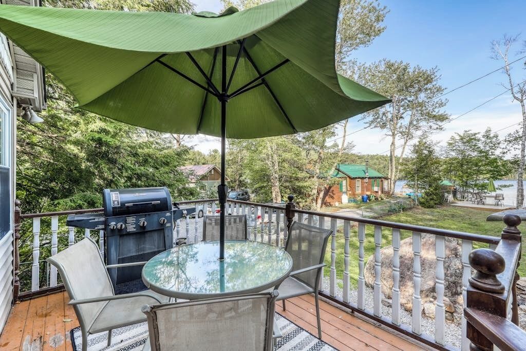 A deck with a glass table and chairs, a grill, and a green umbrella providing shade.