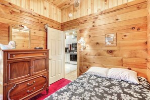 A wooden-paneled room with a bed, a dresser, and a door leading to a kitchen. - Peaceful 3BR Lakefront Adirondacks | Dock (Brant Lake)