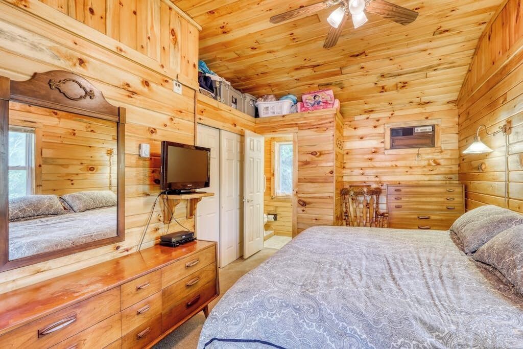 A wooden cabin bedroom with a bed, dresser, television, and a ceiling fan.