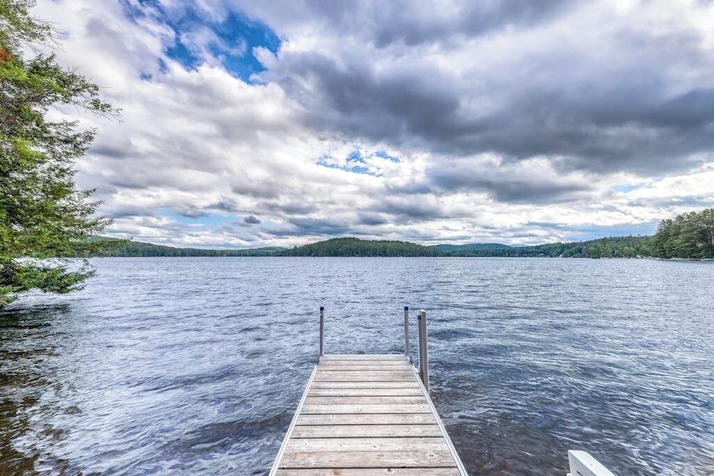 En träbrygga som sträcker sig ut i en sjö, med en skogbeklädd strand och en molnig himmel.