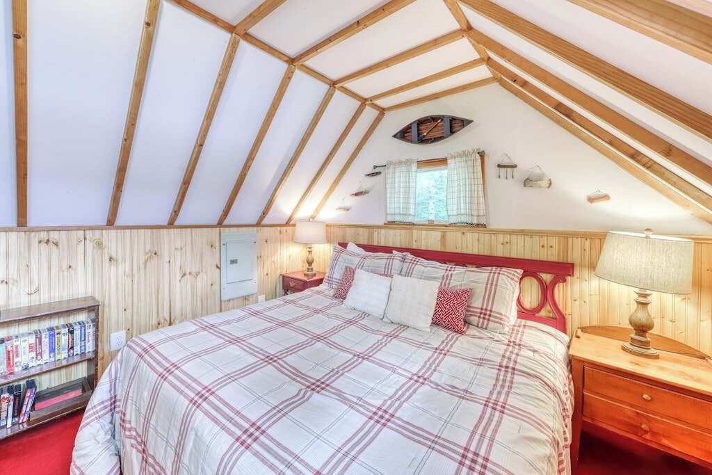 Une chambre avec un plafond en pente, un lit avec une couette à carreaux, une table de chevet avec une lampe et une bibliothèque.