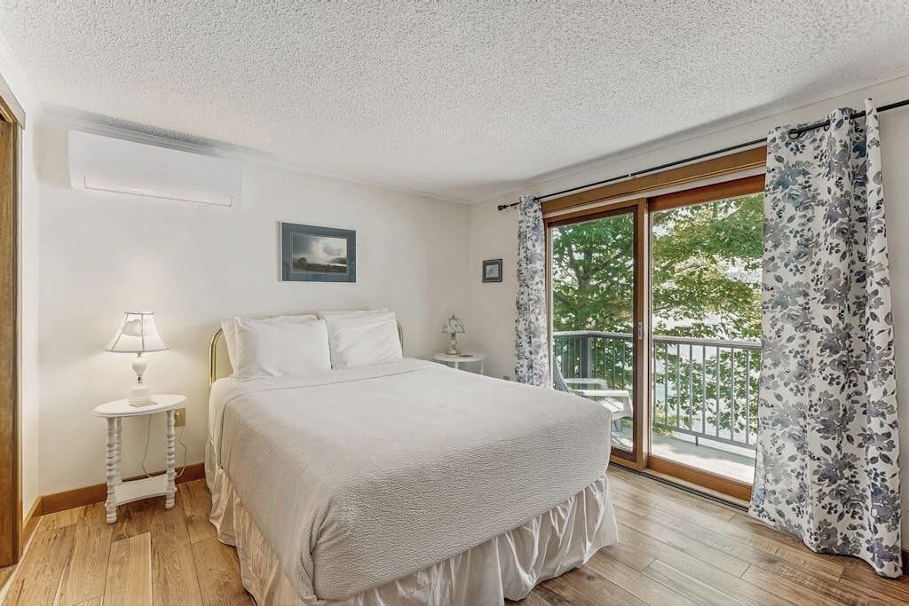 A bedroom with a bed, a nightstand, a lamp, a picture on the wall, and a balcony with curtains.