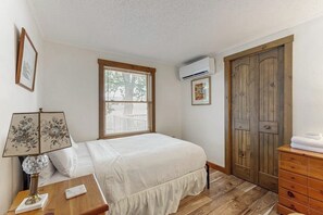 A bedroom with a bed, a wooden dresser, a nightstand with a lamp, a window with blinds, and a wall-mounted air conditioner. - Lakefront Retreat w/Private Dock & Cozy Deck Views (Penn Yan)