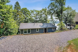A single-story building with a metal roof, surrounded by gravel and trees, with a view of a body of water in the background. - Lakefront Retreat w/Private Dock & Cozy Deck Views (Penn Yan)