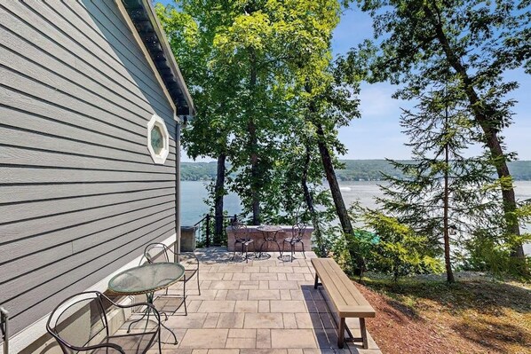 A patio with a table and chairs, overlooking a lake with trees and a building in the background. - Lakefront Retreat w/Private Dock & Cozy Deck Views (Penn Yan)