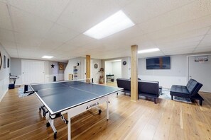 A room with a ping pong table, wooden flooring, a couch, and a basketball hoop. - Relaxing 3BR home with a deck & firepit (Penn Yan)