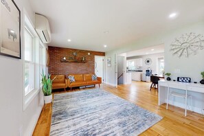 A modern living room with a brick accent wall, a sofa, a rug, and a kitchen in the background. - Relaxing 3BR home with a deck & firepit (Penn Yan)