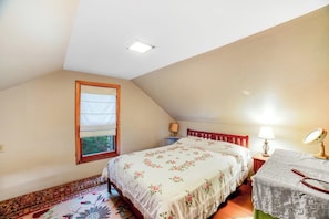 Ein Schlafzimmer mit schrägen Wänden, einem Fenster mit weißer Jalousie, einem Bett mit floralem Bettzeug, einem Nachttisch mit Lampe und einem kleinen Tisch mit Spiegel.