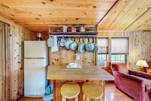 A compact kitchen with a refrigerator, wooden countertop, hanging pots, and a red chair.