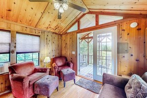 A wooden cabin interior with a ceiling fan, two armchairs, a sofa, and a sliding glass door leading to an outdoor deck.