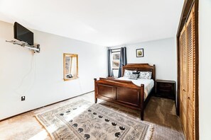 Ein Schlafzimmer mit einem hölzernen Bett, einem an der Wand montierten Fernseher, einem Fenster mit Vorhängen und einem gemusterten Teppich.