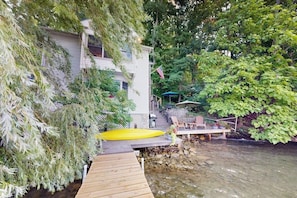 A wooden dock leading to a house with a yellow raft, surrounded by lush greenery and a small deck with chairs. - Willow Bluff | 3 BR lakefront house with private beach & dock (Keuka Park)