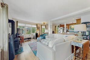 A spacious living room with a sofa, a coffee table, and a fireplace. There is a kitchen area with cabinets and a refrigerator. - Willow Bluff | 3 BR lakefront house with private beach & dock (Keuka Park)