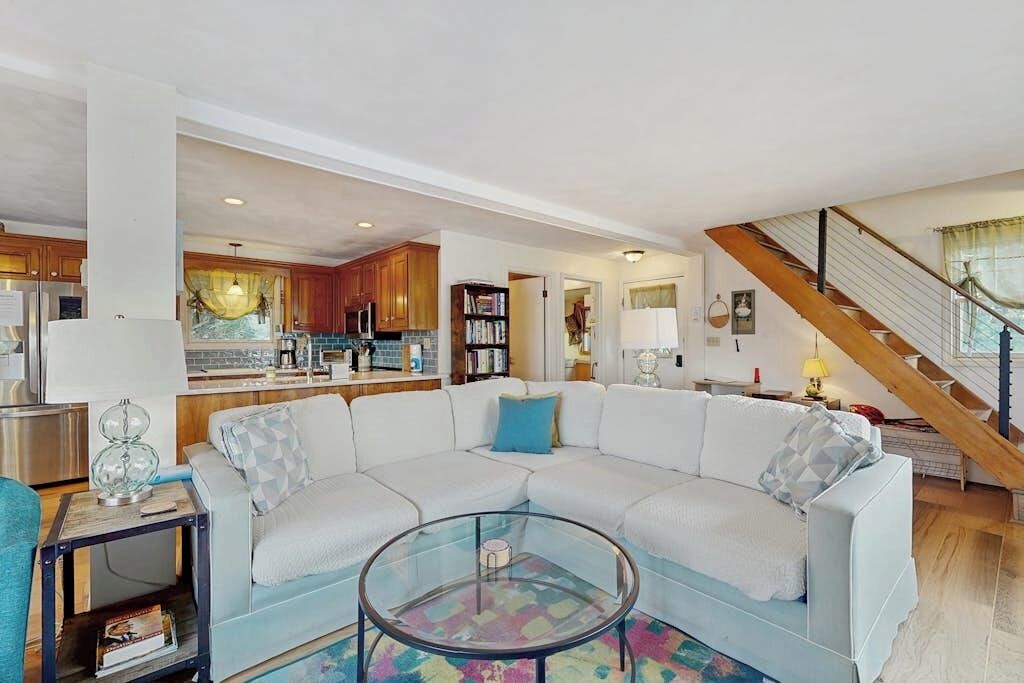 A modern living room with a white sectional sofa, a glass coffee table, and a kitchen in the background.