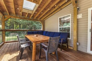 Eine überdachte Terrasse mit einem Holztisch und -stühlen, einem blauen Sofa und einem Fenster mit Blick auf einen Wald.