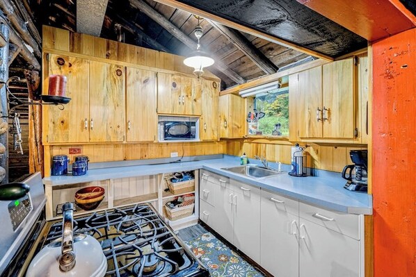 A rustic kitchen with wooden cabinets, a gas stove, and a window overlooking greenery.