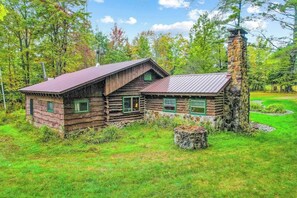 A rustic log cabin with a stone chimney, surrounded by a grassy area and trees.