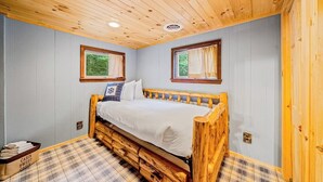 Ein schlicht eingerichtetes Schlafzimmer im Blockhausstil mit einem hölzernen Bett, einem karierten Boden und zwei mit Vorhängen versehenen Fenstern.