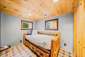 Quarto em estilo cabana com cama de madeira, piso xadrez e duas janelas com cortinas.
