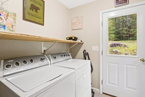 A laundry area with a front-loading washer and dryer, a shelf with items, and a door leading to a forested area.