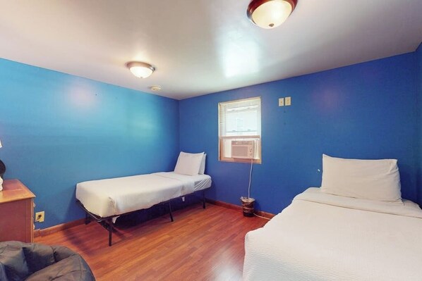 A room with two single beds, a wooden floor, a window with blinds, and a ceiling light fixture.