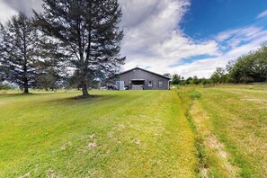 A single-story building with a garage, surrounded by a grassy field and trees. - Italy Hill Estate | Charming Retreat 2BR Finger Lakes - Hot Tub (Branchport)