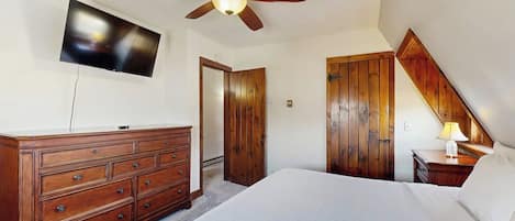 A bedroom with a bed, wooden dresser, ceiling fan, TV, and a lamp.