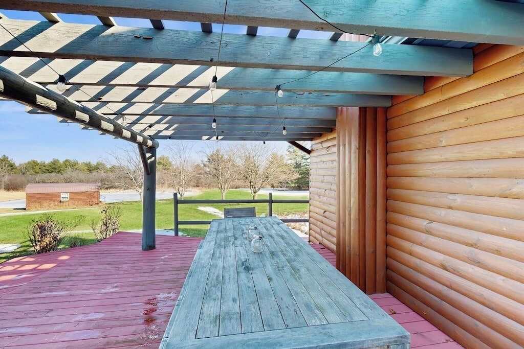 Un terrazzo in legno con tavolo da picnic, un pergolato con luci a stringa e vista su un paesaggio rurale.