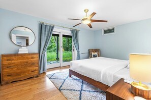 Ein Schlafzimmer mit einem Bett, einem hölzernen Schminktisch, einem Deckenventilator und Blick ins Grüne.