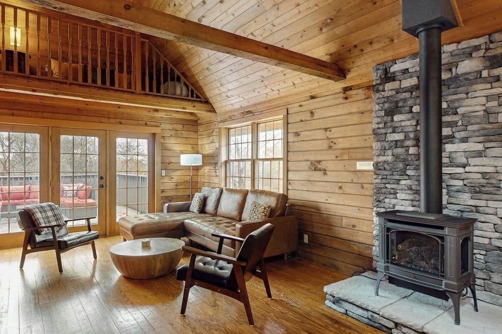 A log cabin interior with a stone fireplace, wooden furniture, and a loft area.