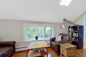 A living room with a brown leather sofa, a wooden coffee table, a small side table, a bookshelf, and a large window with a view of trees. - 3BR mountain-view house with fireplace (Thurman)
