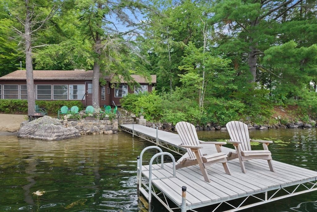 A wooden dock with two Adirondack chairs, a stone structure, and a building with windows in the background.