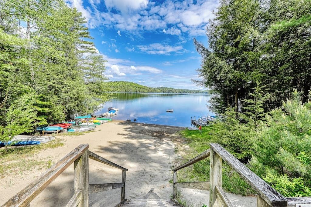 A wooden staircase leading to a sandy beach with boats docked on the left, surrounded by dense trees and a calm lake under a partly cloudy sky.