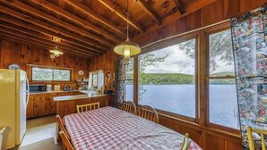 Une cabane avec un intérieur en bois, une table à manger recouverte d’une nappe à carreaux, un coin cuisine et une grande fenêtre donnant sur un lac.