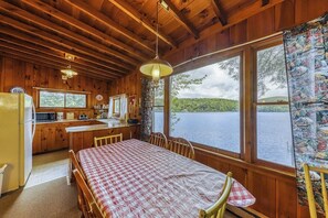 Een chalet met houten inrichting, een eettafel met geruit tafelkleed, een keukenhoek en een groot raam met uitzicht op een meer.