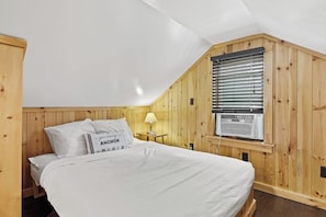Una camera da letto con soffitto in legno spiovente, un letto con biancheria bianca, un comodino con una lampada e una finestra con persiane.