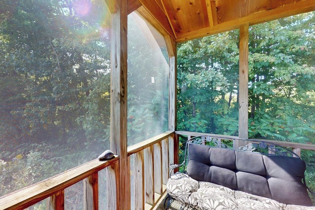 Una terraza de madera con zona de estar acolchada y grandes ventanales con vistas a un área boscosa.