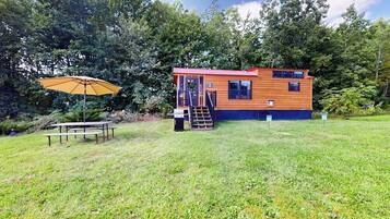 Une cabane en bois avec un toit rouge, une table de pique-nique et un parasol, située dans un espace verdoyant entouré d’arbres.