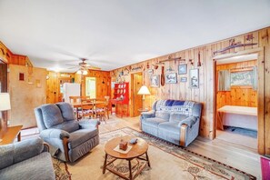A cozy living room with wooden walls, a ceiling fan, a sofa, a coffee table, and a dining area with a red table and chairs. - 3BR Lakefront - Fireplace - Dock - Screened Porch (Chestertown)