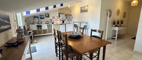 A dining area with a wooden table and chairs, a kitchen in the background, and a decorative wall with a clock.
